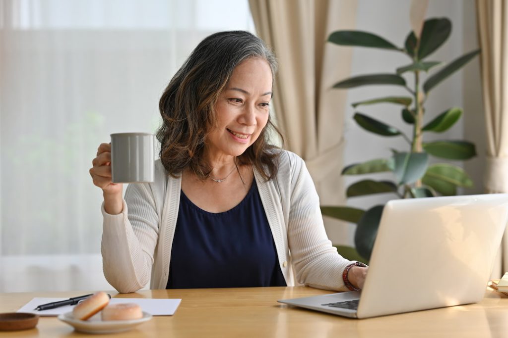 Woman with grey hair enjoying a coffee tasting experience virtually with coworkers, sitting at a desk in front of a laptop