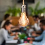 A rustic lightbulb in the foreground in front of several employees participating in a creativity brainstorming session
