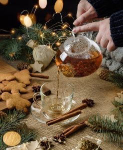 Someone pouring holiday tea into a mug from a glass teapot surrounded by holiday lights, gingerbread cookies, and festive pine garlands