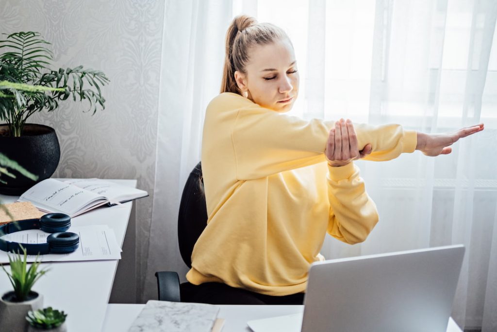 Woman stretching at her desk while working from home in a well lit room surrounded by office plants