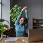 Female employee at her desk working from home stretching surrounded by office plants