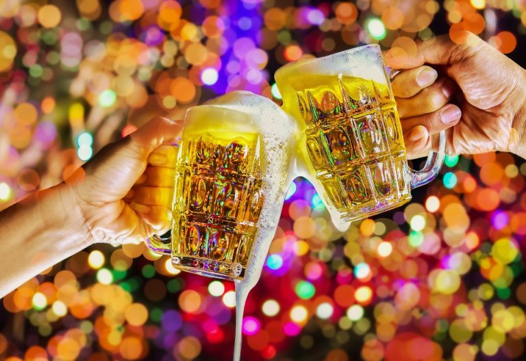 Two people toasting with full glasses of beer with festive holiday lights in the background