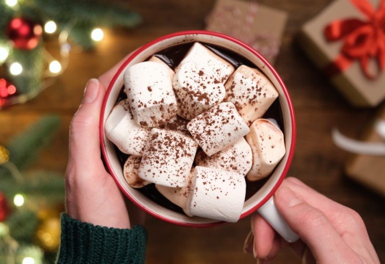 A steaming mug of hot chocolate topped with large marshmallows, with holiday gifts in the background