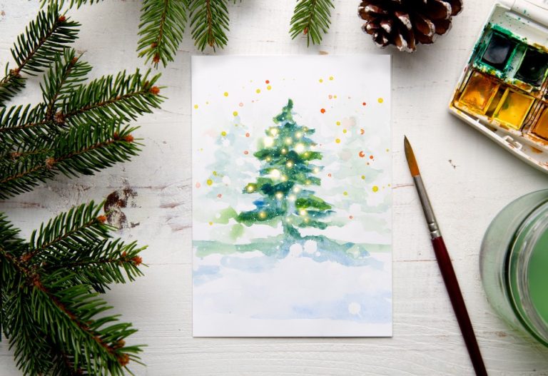 A paintbrush resting near a painting of a green holiday tree, surrounded by pine decorations