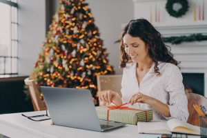 A female employee opens a gift at her desk with a festive tree in the background of her remote office.