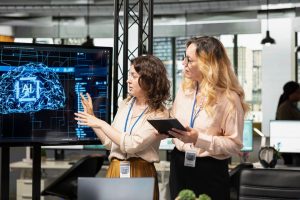 Two employees in an office talking together and standing next to a tv screen that says "AI."